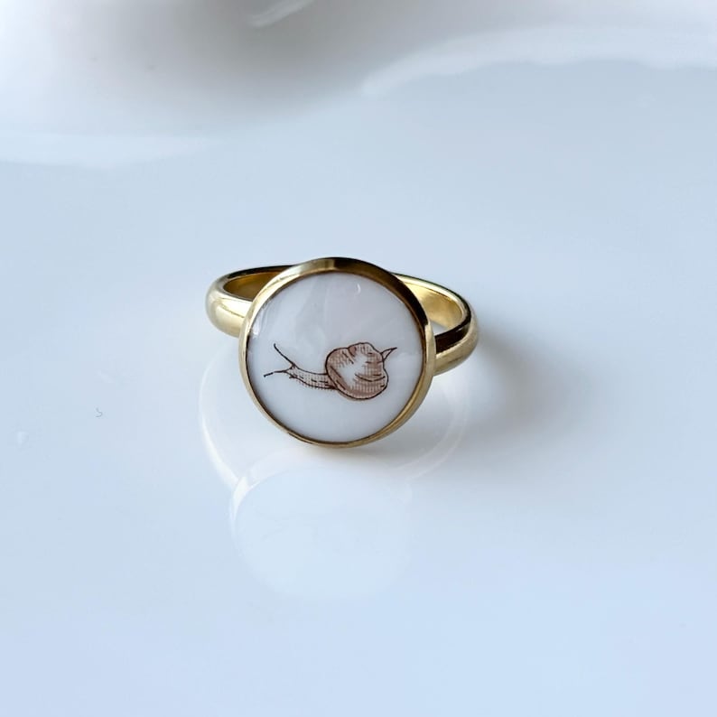 May include: A gold-colored ring with a round, white face featuring a brown snail illustration. The ring has a polished finish and is set against a white background. The snail is drawn in a simple, illustrative style.