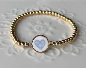Puede incluir: Pulsera dorada de cuentas con un dije redondo con un diseño de corazón azul claro sobre fondo blanco. El dije está enmarcado en oro. La pulsera está sobre una superficie blanca texturizada.
