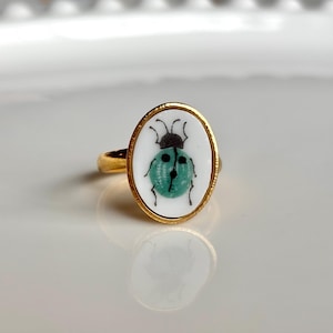May include: Gold-toned ring with an oval white face featuring a teal ladybug design. The ladybug has a black head and black details. The ring is set against a white background.