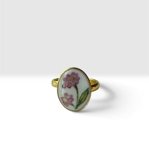 May include: A gold-toned ring featuring an oval-shaped white ceramic inlay. The inlay is decorated with a hand-painted design of two small pink flowers with green leaves and stems. The metal band is smooth and circular, framing the floral centrepiece.
