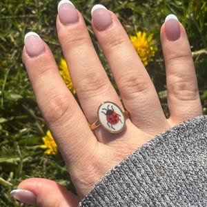 May include: Gold ring with an oval, white enamel face displaying a red ladybird design. The ring is worn on a hand with manicured nails. The background is blurred, showing green grass and yellow flowers.