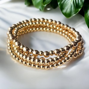 May include: Three gold beaded bracelets stacked on top of each other. The bracelets are made of small, round beads.