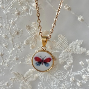 May include: A gold-coloured chain necklace with a round pendant. The pendant has a white background with a butterfly design in red and blue. The butterfly is within a gold-coloured frame.
