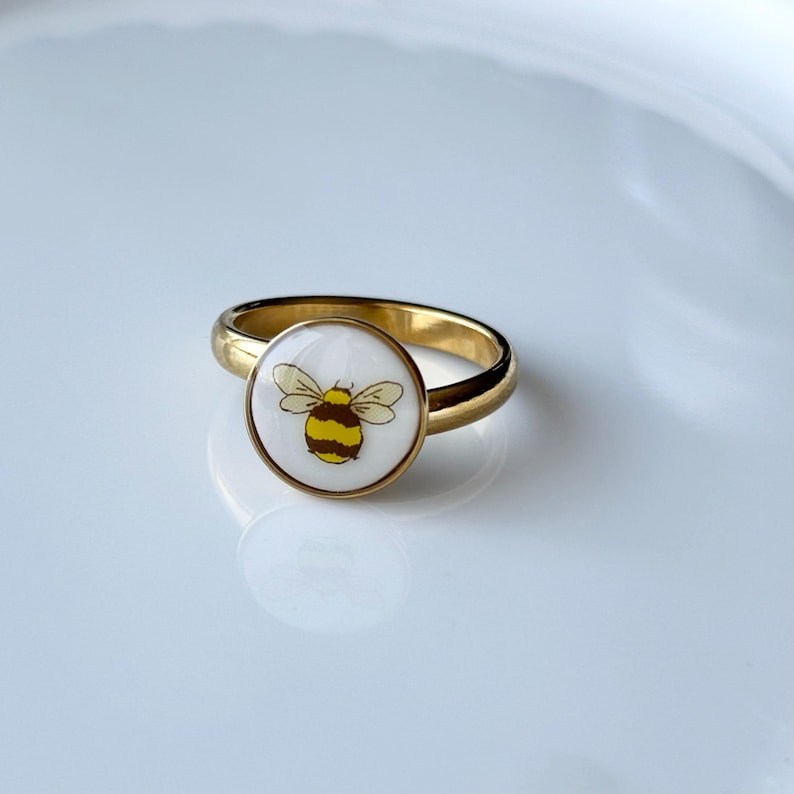 May include: A gold-colored ring with a circular white face featuring a yellow and black bee illustration. The ring is set against a white background, highlighting the jewelry's design and details. The ring is a piece of fashion jewelry.