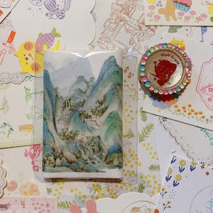 May include: A collection of colorful patterned paper sheets with a watercolor painting of a mountain landscape. The paper sheets have various designs including flowers, houses, and animals. There is a small pink and gold decorative seal with the text "Whim Craft" on it.