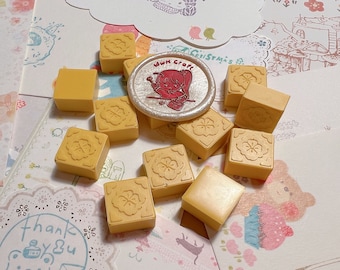 May include: A collection of 10 small, square, yellow rubber stamps with a floral design. The stamps are arranged in a semi-circle around a round, silver wax seal with a red and white illustration of a girl holding a paintbrush. The wax seal has the text "Whim Craft" on it.