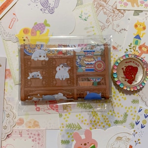 May include: A small, brown, wooden box with a clear plastic cover. The box has a decorative design with a repeating pattern of cats and flowers. The box is sitting on a table with a variety of colorful papers and stickers.