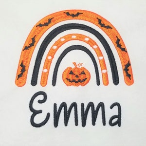 May include: A black and orange rainbow with bats and white dots, a jack-o'-lantern in the center, and the name "Emma" underneath.