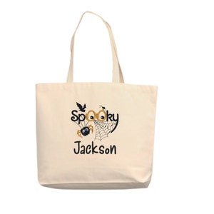 May include: A white canvas tote bag with a black and orange Halloween design. The design features a spider web, a spider, and the words "Spooky Jackson".