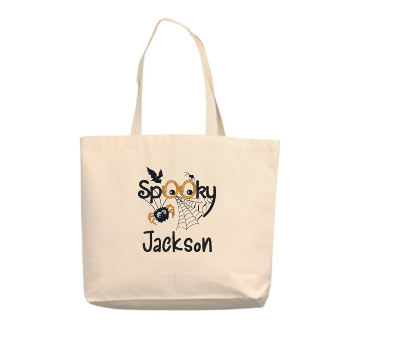 May include: A white canvas tote bag with a black and orange Halloween design. The design features a spider web, a spider, and the words "Spooky Jackson".