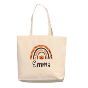 May include: A white canvas tote bag with an orange, black, and white rainbow design. The rainbow has bats and a pumpkin in the center. The name "Emma" is written below the rainbow.
