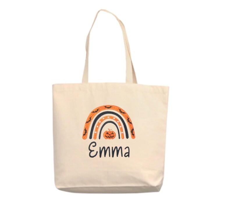 May include: A white canvas tote bag with an orange, black, and white rainbow design. The rainbow has bats and a pumpkin in the center. The name "Emma" is written below the rainbow.