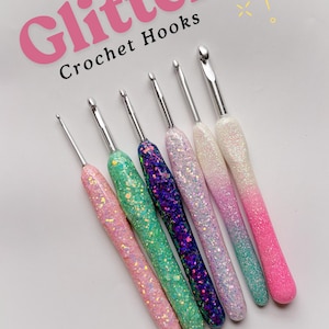 Könnte beinhalten: Eine Sammlung von sechs Häkelnadeln mit Glitzergriffen in verschiedenen Farben, darunter Rosa, Grün, Lila und Weiß. Der Text "Glitter Crochet Hooks" ist in Rosa und Schwarz dargestellt. Die Häkelnadeln sind auf weißem Hintergrund angeordnet.