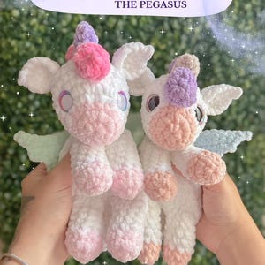 May include: Two crocheted unicorn plushies, one with pink and white accents and the other with purple and white accents. The plushies have wings and are held in a person's hands. The text "Pearl The Pegasus crochet pattern" is at the bottom of the image.