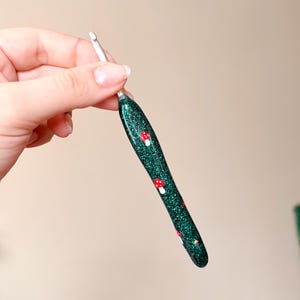 Op de afbeelding: Een groene glitter haaknaald met rode en witte paddenstoelontwerpen. De haak heeft een zilveren metalen punt en een afgeronde handgreep. De handgreep is versierd met kleine rode en witte paddenstoelen.
