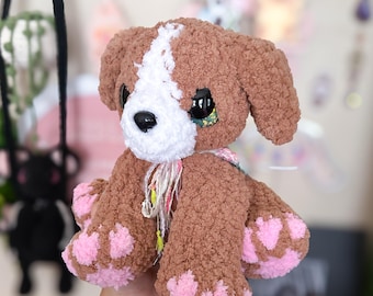Puede incluir: Un cachorro de peluche marrón y blanco hecho a mano con almohadillas rosas. El cachorro tiene ojos negros grandes y una raya blanca en la cabeza. Es un juguete suave y tierno.