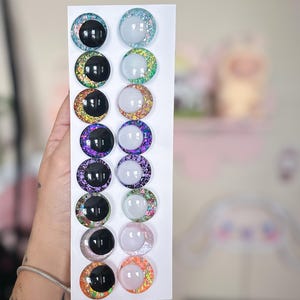 May include: A collection of twelve colorful, round craft eyes with glitter accents. The eyes are arranged in two columns on a white card. The eyes feature a variety of colors, including black, blue, purple, and orange, with glitter rims and reflective centers.