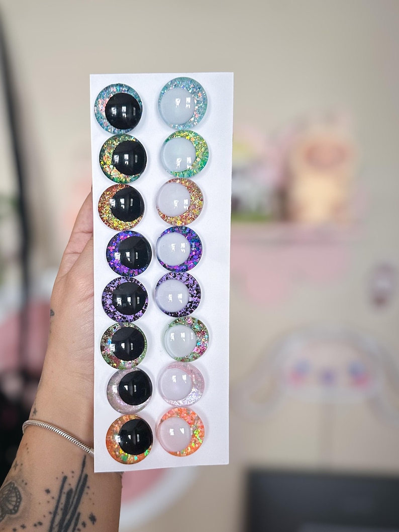 May include: A collection of twelve colorful, round craft eyes with glitter accents. The eyes are arranged in two columns on a white card. The eyes feature a variety of colors, including black, blue, purple, and orange, with glitter rims and reflective centers.