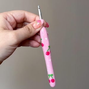 May include: A pink crochet hook with a cherry pattern and glitter accents. The hook features a silver metal tip and a rounded handle decorated with red cherries and green leaves. The handle also has white star-shaped accents.