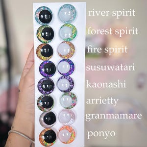 May include: A white card displays twelve round, glittery cabochons in various colors, including black, blue, green, purple, and orange. Each cabochon has a black or white center. Text on the right side of the card reads "river spirit", "forest spirit", and other names.