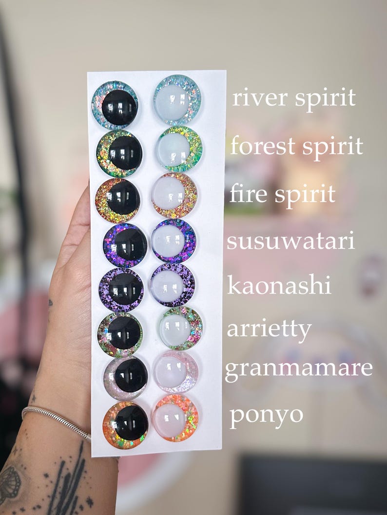 May include: A white card displays twelve round, glittery cabochons in various colors, including black, blue, green, purple, and orange. Each cabochon has a black or white center. Text on the right side of the card reads "river spirit", "forest spirit", and other names.