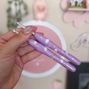 May include: Two crochet hooks with lavender glitter handles. The handles are decorated with white clouds, gold stars, and silver glitter. The hooks are held in a hand, with a blurred background of decorative items.