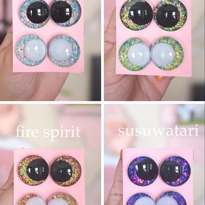 May include: Four sets of decorative glass eyes on pink cards, each labeled with a different theme: "river spirit," "forest spirit," "fire spirit," and "susuwatari." The eyes have black or white centers with glittery, colorful rims.