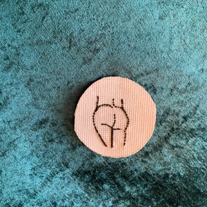Puede incluir: Un parche redondo beige con bordado negro de una figura humana estilizada. La figura está delineada con hilo negro y tiene una forma de cruz en el centro.