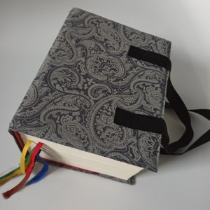 Può includere: Una copertina per libro grigia con motivo paisley, cinturini neri e dorso rosso. La copertina ha un dorso da libro ed è realizzata in tessuto.