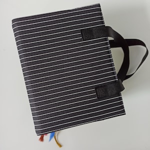 Op de afbeelding: Een zwart-wit gestreept notitieboek met een zwart handvat. Het notitieboek heeft een stoffen kaft met dunne witte horizontale strepen op een zwarte achtergrond. Er zijn kleurrijke lint-bladwijzers zichtbaar.