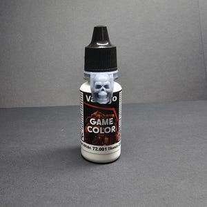 Może przedstawiać: Butelka białej farby Vallejo Game Color z czarną nakrętką i jasnoniebieskim uchwytem na nakrętkę w kształcie czaszki. Etykieta głosi "Game Color" i "White 72.001 Blanco".