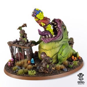 Op de afbeelding: Een geschilderd miniatuurdiorama met een groot groen monster met een wijd open mond, een kleiner groen wezen dat de lucht in wordt gelanceerd en een groen figuur op een houten kooi. De basis bevat kleine paddenstoelen en de tekst GORK'S SCULPTS.