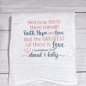 May include: White quilted blanket with embroidered text in pink and blue. The text reads "And now these three remain Faith, Hope and Love But the GREATEST of these is Love" with a heart symbol and "1 Corinthians 13:13 david & katy".
