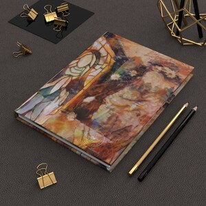 May include: A hardcover journal with an abstract art design in warm tones of brown, orange, and yellow. The journal is surrounded by gold and black office supplies, including paper clips and pencils. The journal has the text "Find Your Soul Me" on the cover.
