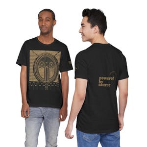 Puede incluir: Camisetas negras con un diseño gráfico dorado en la parte delantera y el texto "powered by source" en la parte trasera. El diseño frontal es un patrón circular que se asemeja a una cara.