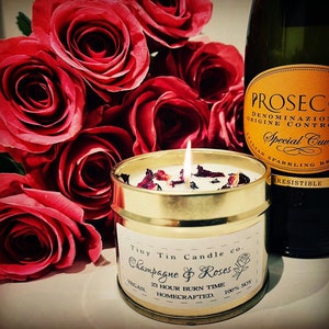 Celebrate with Tiny Tin Candle Co.'s 'Champagne & Roses' homemade soy candle, lit beside a bouquet of red roses and a bottle of Prosecco, setting a romantic and festive mood