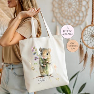May include: A cream-coloured canvas tote bag featuring a watercolour illustration of a small brown mouse holding a sprig of purple flowers. Below the mouse, the name Chloe is printed in black script. Two circular badges state 8 DESIGNS AVAILABLE and ADD YOUR OWN TEXT.