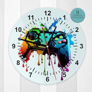 May include: A round wall clock featuring a colourful graphic of a video game controller with paint drip effects in rainbow hues. The clock face has black numbers from 1 to 12, black hour and minute hands, and a red second hand. A circular badge reads 8 DESIGNS AVAILABLE.