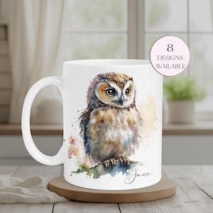 May include: White ceramic mug with a watercolour illustration of an owl. The owl is depicted with brown, tan, and blue feathers. The mug has the cursive text "Jane" and a pink circle with the text "8 DESIGNS AVAILABLE".