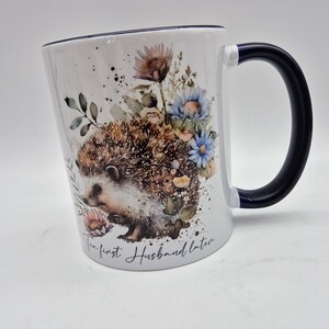 May include: White ceramic mug with a black handle and rim. The mug features a watercolour illustration of a hedgehog surrounded by flowers in shades of blue, pink, and brown. The text "The first Husband later" is written on the mug.