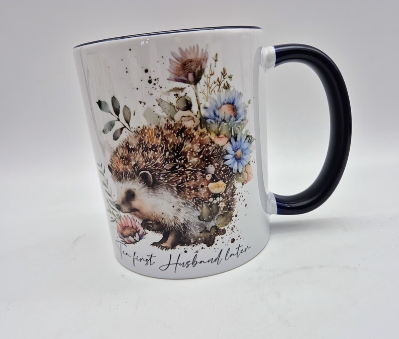 May include: White ceramic mug with a black handle and rim. The mug features a watercolour illustration of a hedgehog surrounded by flowers in shades of blue, pink, and brown. The text "The first Husband later" is written on the mug.