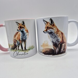 May include: Two white ceramic mugs with fox illustrations. One mug has a pink handle and the name "Charlie" written on it. The other mug has a white handle. The foxes are depicted with orange, brown, and white fur.