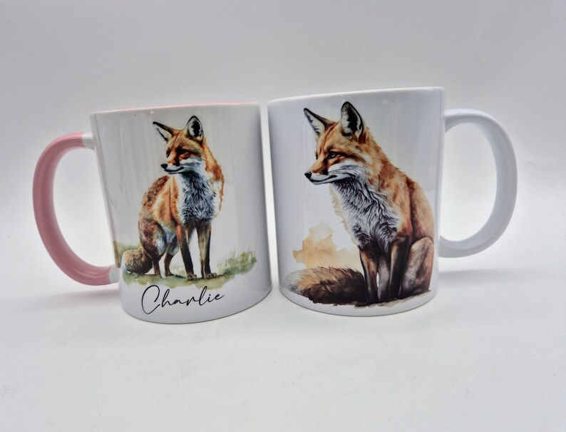 May include: Two white ceramic mugs with fox illustrations. One mug has a pink handle and the name "Charlie" written on it. The other mug has a white handle. The foxes are depicted with orange, brown, and white fur.