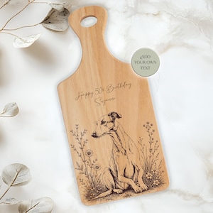 May include: Wooden cutting board with a handle, engraved with a dog illustration and the text "Happy 50th Birthday Suzanne". The board features a light brown color and a natural wood grain.