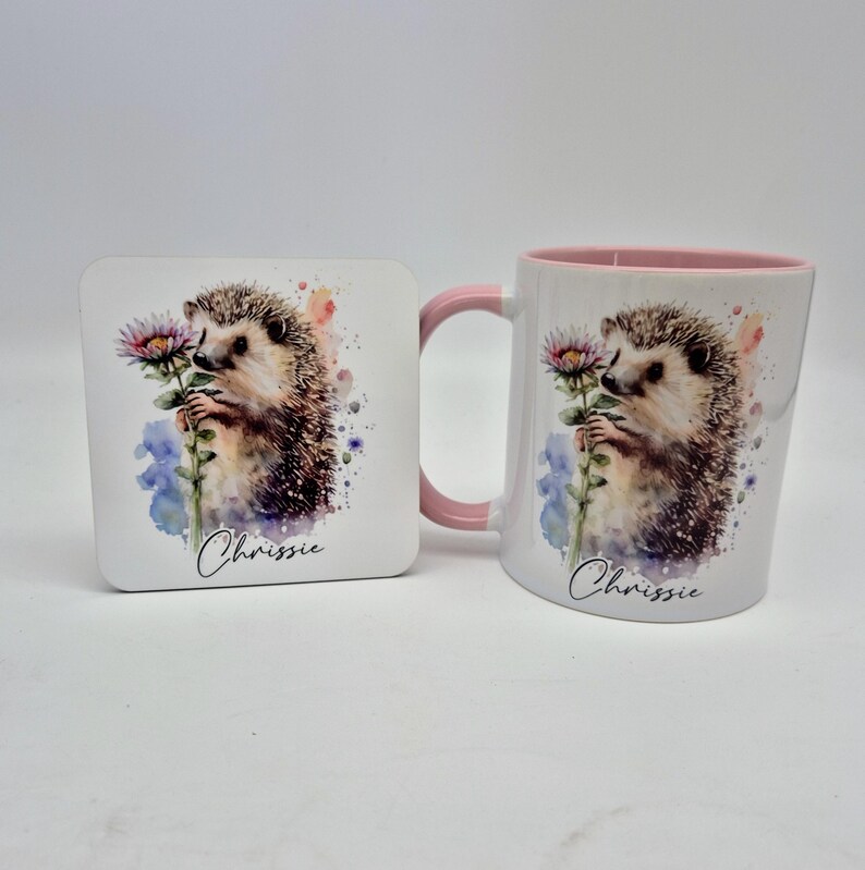 May include: A white ceramic mug and coaster set featuring a watercolour hedgehog holding a flower. The mug has a pink handle and interior. The coaster and mug are printed with the name "Chrissie".
