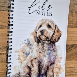 May include: A spiral-bound notebook with a watercolour illustration of a light brown dog. The cover reads "Lil's NOTES" in script. The notebook has lined pages and a white cover, perfect for jotting down notes.