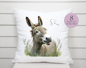 May include: White decorative pillow featuring a watercolor illustration of a donkey. The donkey is light brown and gray, with a smiling expression. The pillow has the name "Sam" and a pink circle with "8 DESIGNS AVAILABLE".