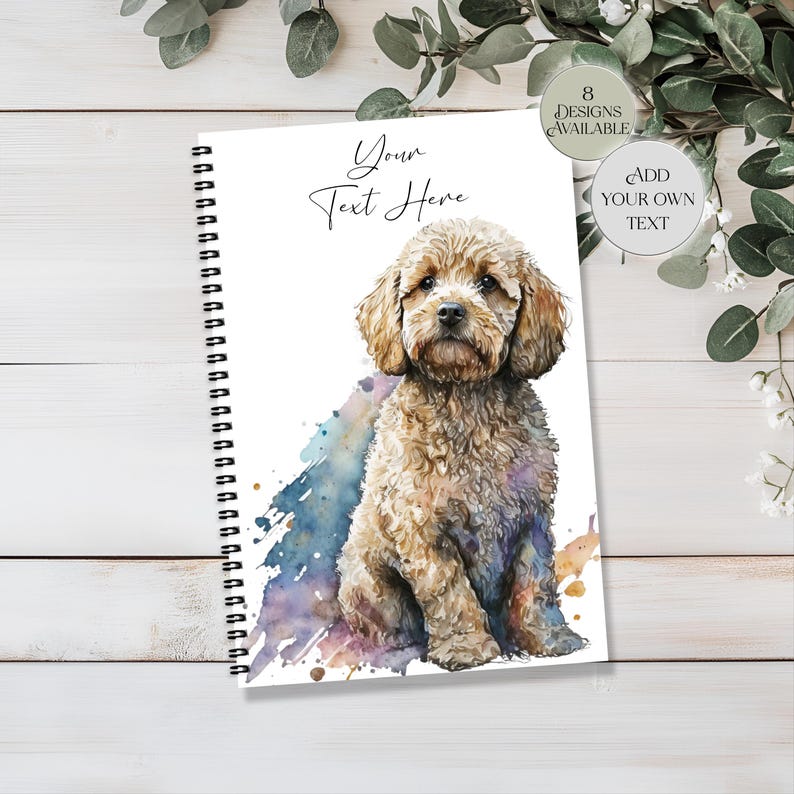 May include: A spiral-bound notebook with a watercolour illustration of a light brown dog. The notebook features the text "Your Text Here" and a watercolour splash in blue, purple, and gold. Additional text indicates "8 Designs Available" and "Add Your Own Text".