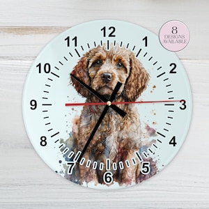 May include: A round wall clock featuring a watercolor painting of a brown dog. The clock face has black numerals and hands, with a red second hand. The text "8 DESIGNS AVAILABLE" is in a pink circle.