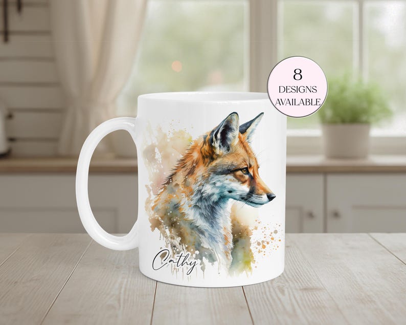 May include: White ceramic mug featuring a watercolor illustration of a fox with orange, brown, and blue hues. The mug has a white handle and the name "Cathy" is written in script. A pink circle indicates "8 designs available".